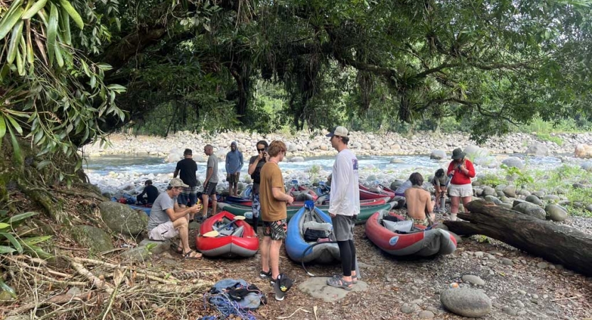 On the bank of a river, a group of students stand and sit near beached watercraft. 